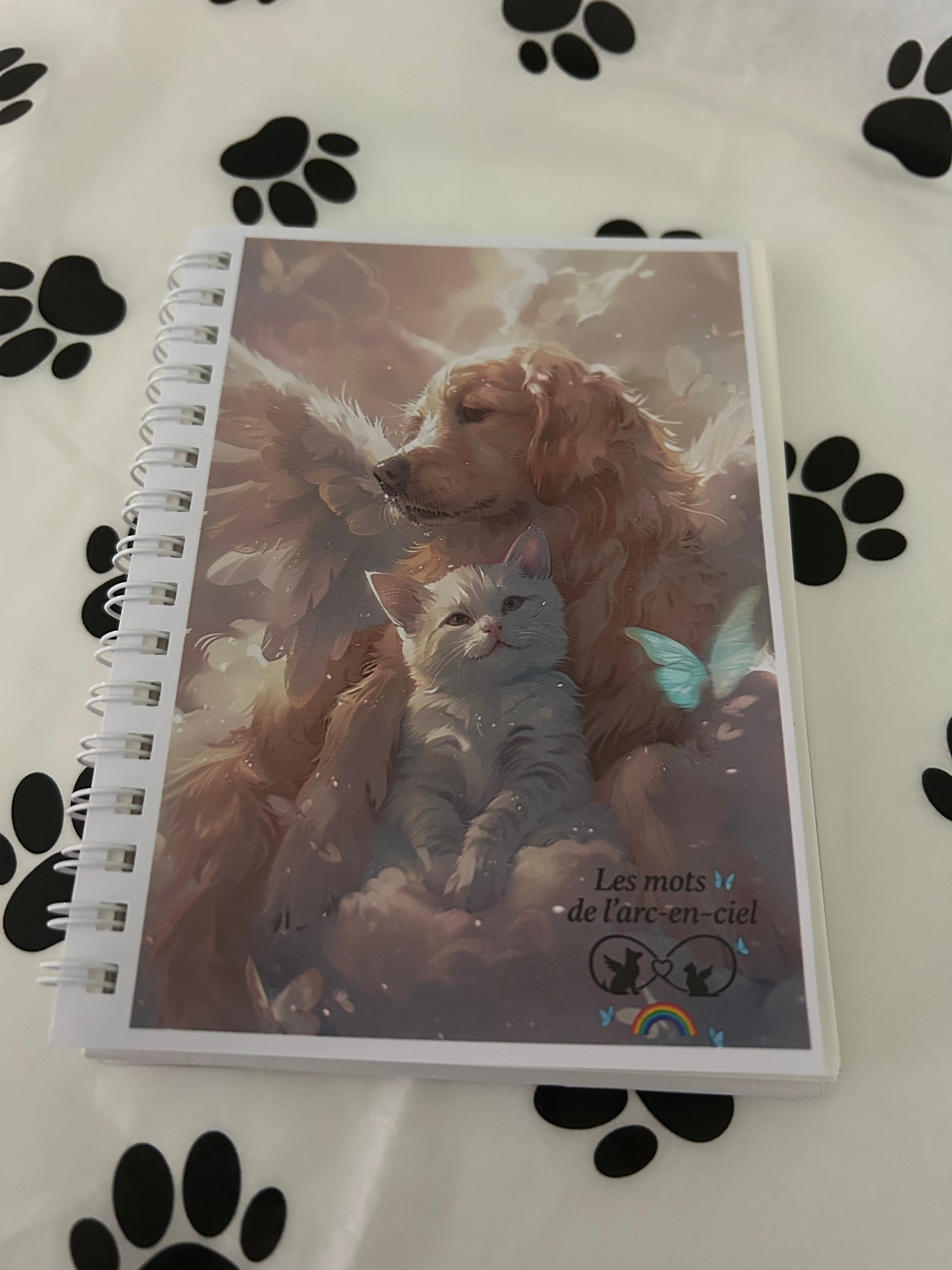 Carnet à lignes pour écrire les signes, pensées et souvenirs après la perte d’un animal, avec une couverture illustrée d’un chien et d’un chat dans les nuages, entourés de lumière et d’un papillon bleu. Cahier Les Mots de l’Arc-en-Ciel posé sur un papier décoré d’empreintes de pattes.