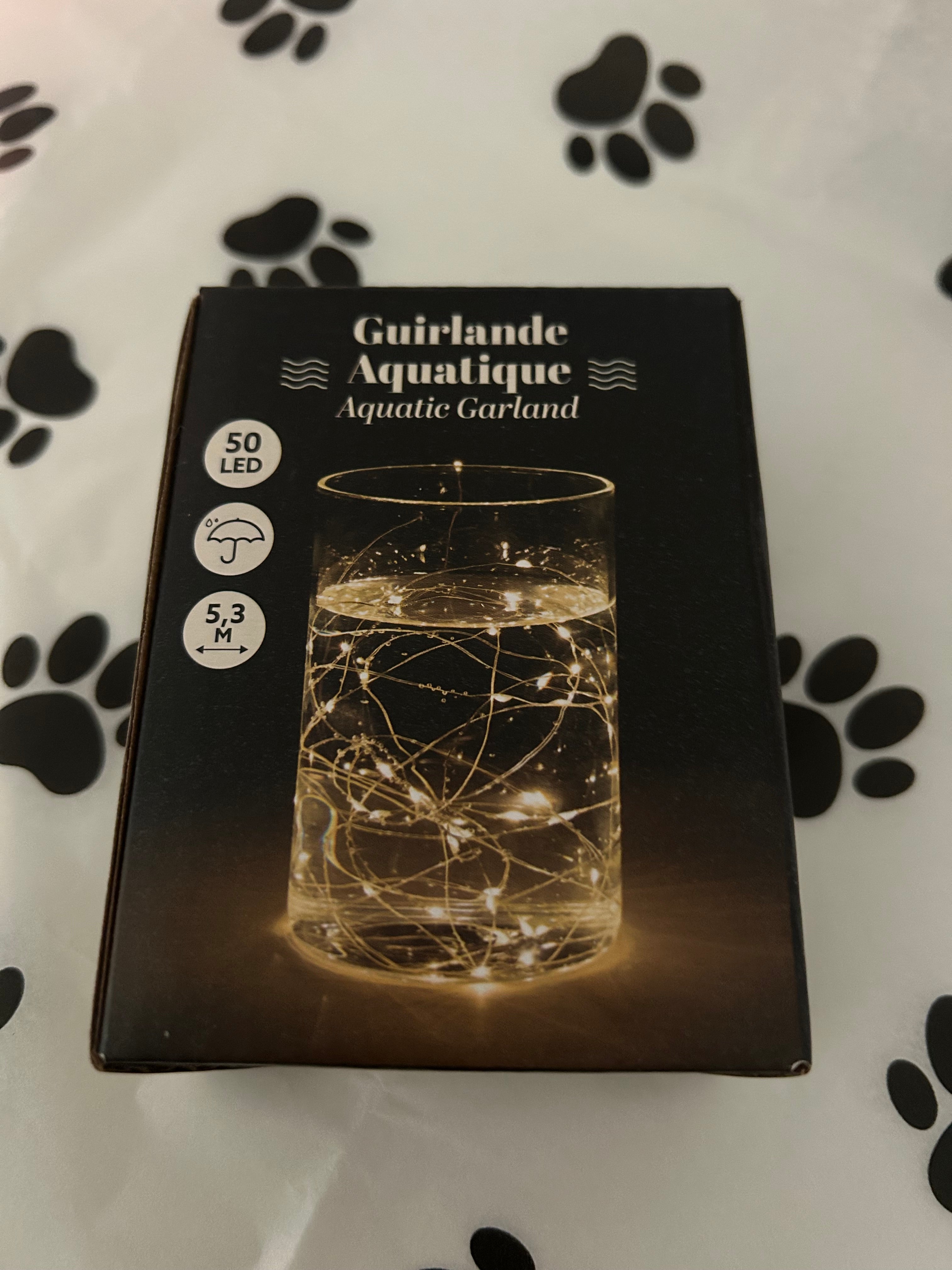 Boîte de guirlande lumineuse aquatique LED, 5,3 mètres, lumière dorée chaude, posée sur un papier décoré d’empreintes de pattes. Accessoire lumineux utilisé dans le coffret du deuil animalier pour créer une ambiance douce et réconfortante.