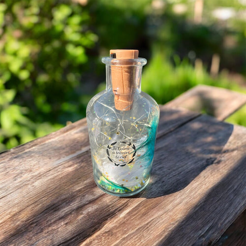 Bouteille lumineuse souvenir posée sur une table en bois au soleil, remplie de petites lumières et décorée d’un message symbolique — création destinée à honorer la mémoire d’un animal.