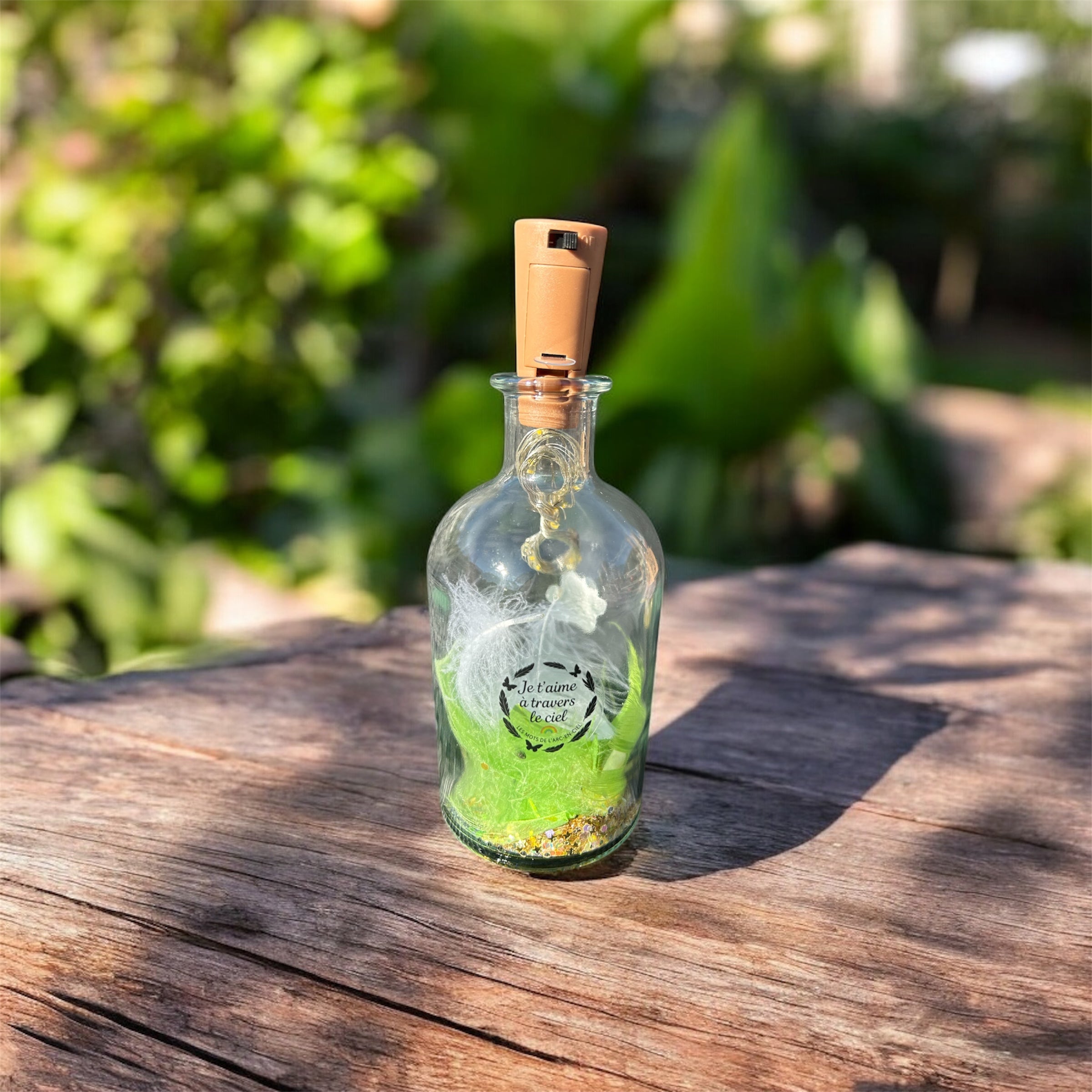 Bouteille lumineuse souvenir posée sur une table en bois au soleil, remplie de petites lumières et décorée d’un message symbolique — création destinée à honorer la mémoire d’un animal.
